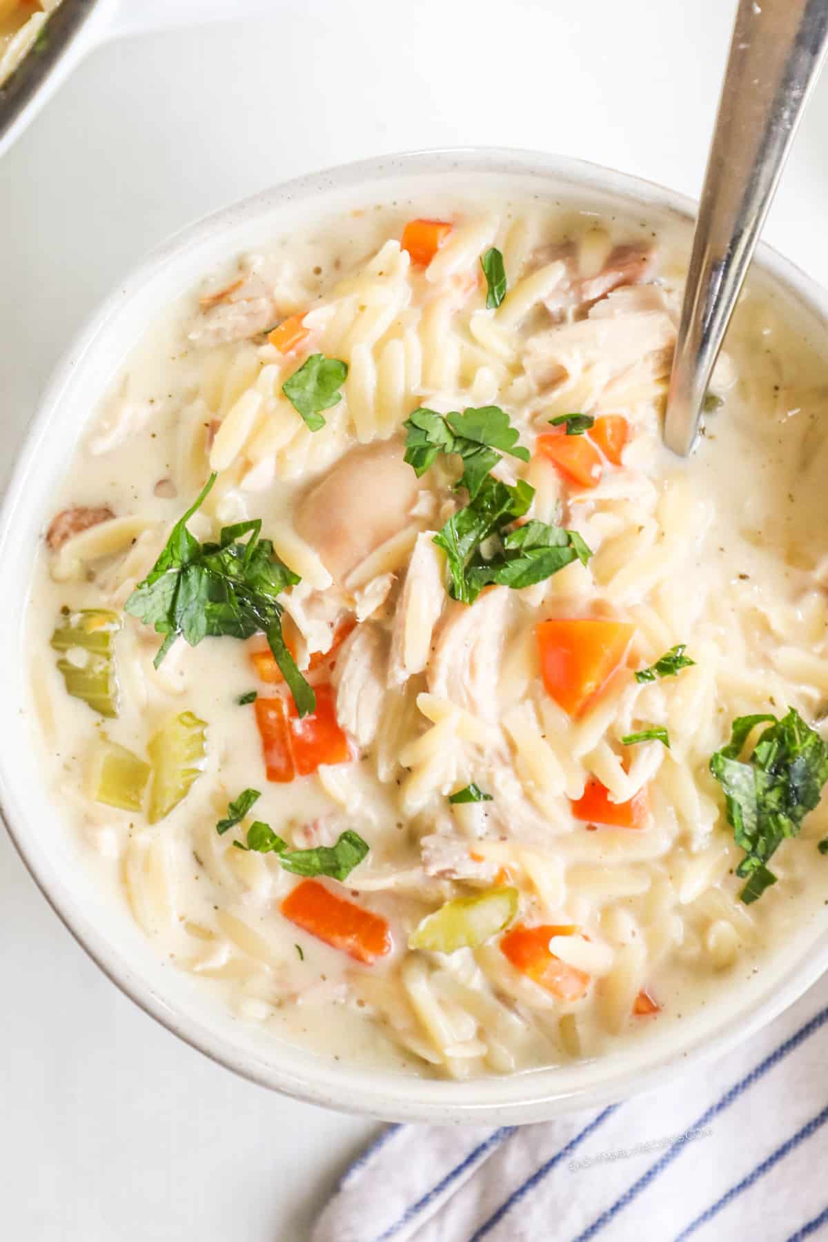 A white bowl full of creamy lemon chicken orzo soup with a silver spoon in it.