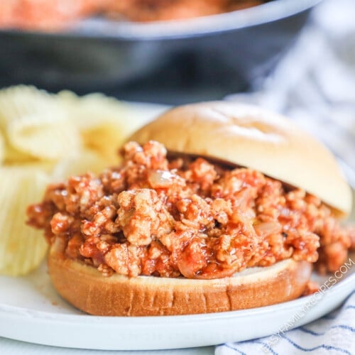 a bun on a plate piled with sloppy joe filling with a side of potato chips.