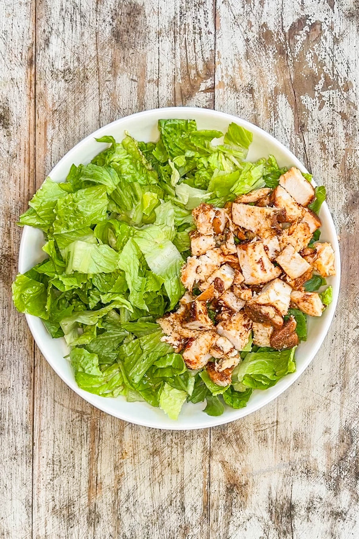 Step 1 of making BBQ Chicken Ranch Salad. Green lettuce and chicken are in a white bowl on a distressed wood background.