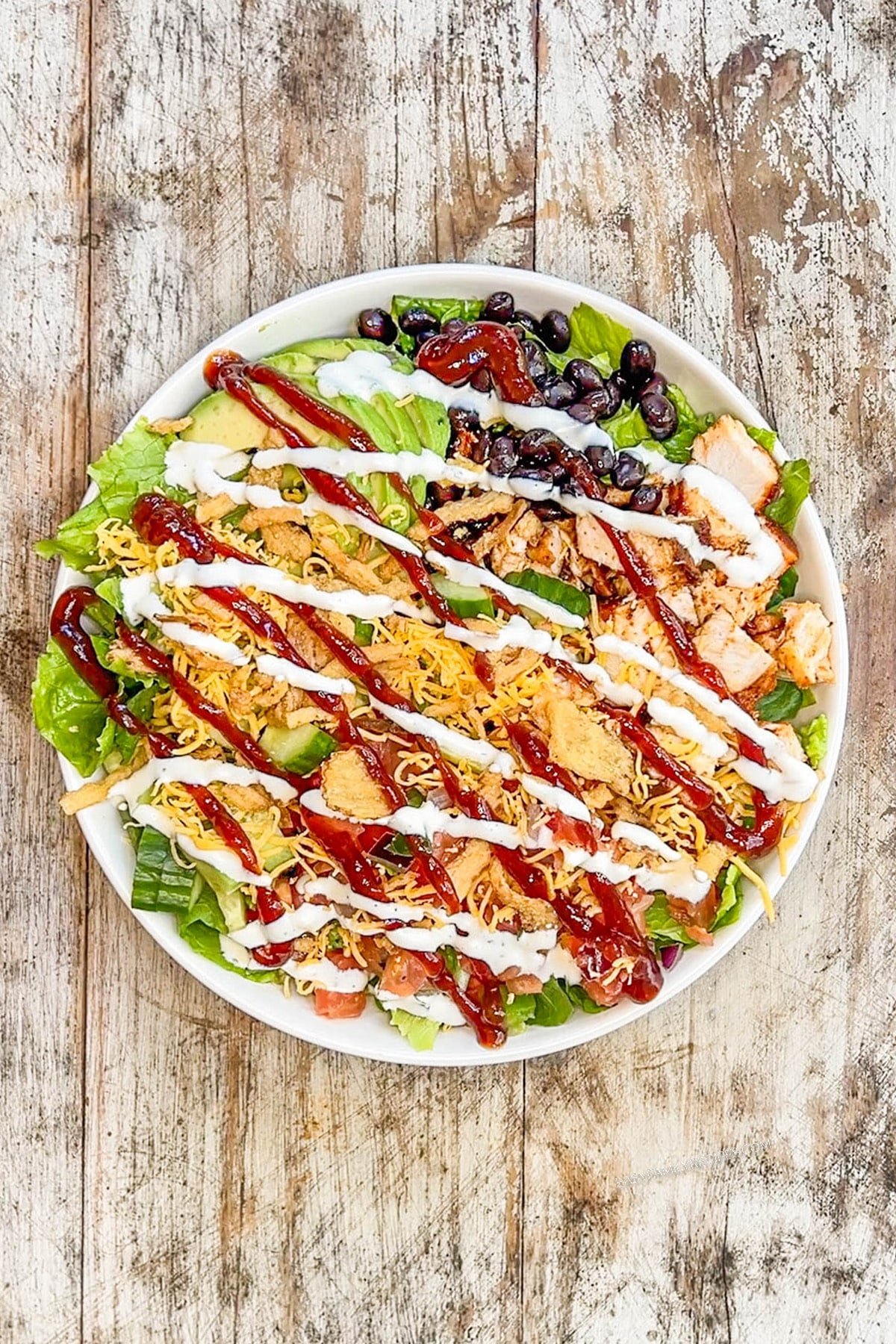 Step 4 of making BBQ Chicken Ranch Salad. BBQ sauce and ranch dressing is drizzled over a salad in a white bowl on a distressed wood background.