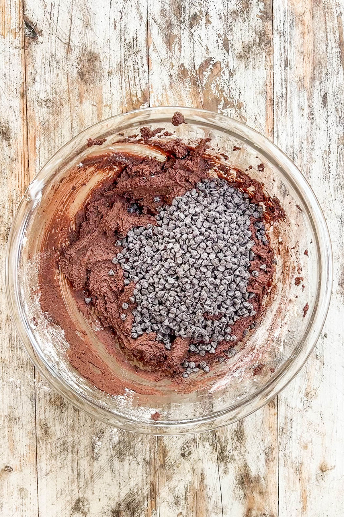 Step 4 for making chocolate marshmallow cookies: Chocolate chips are mixed into the dough in a glass bowl on a distressed wood background.