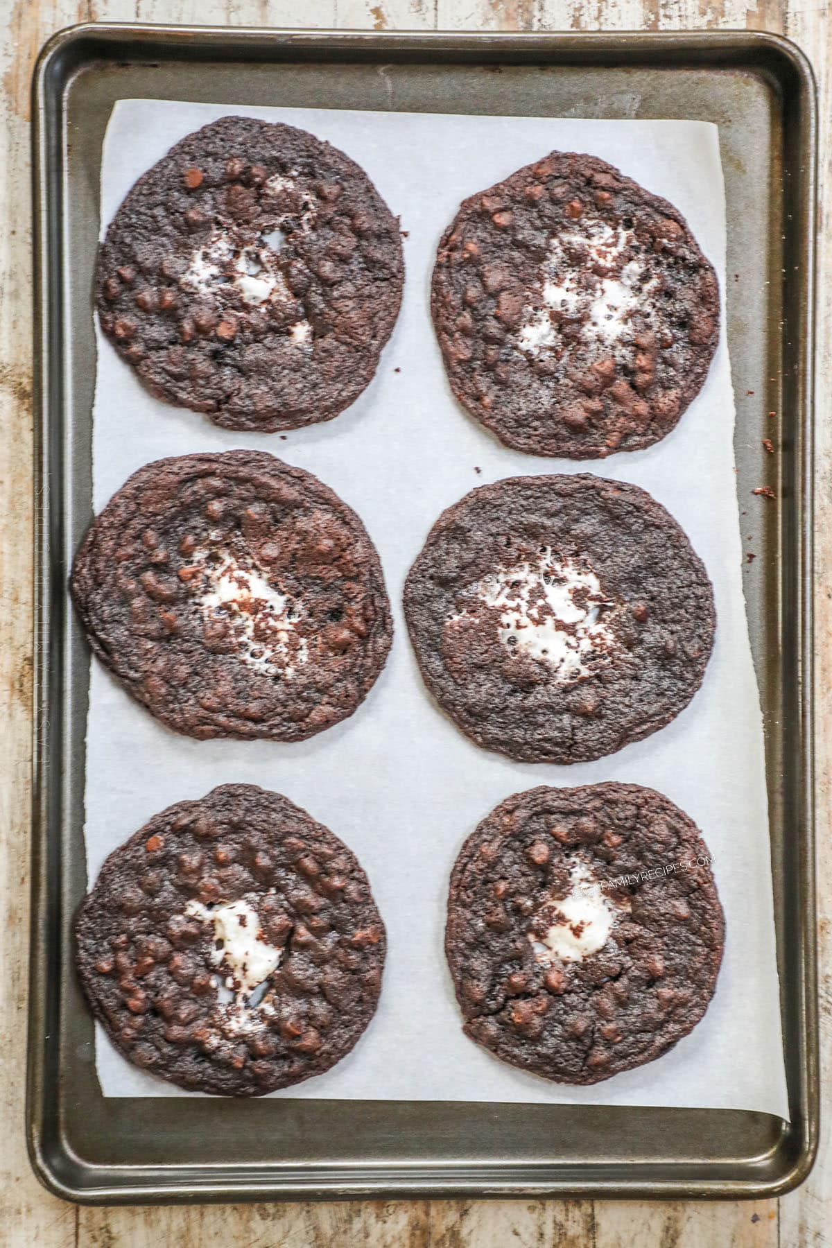 A top view of a dark silver baking sheet lined with parchment paper, and six chocolate marshmallow cookies.