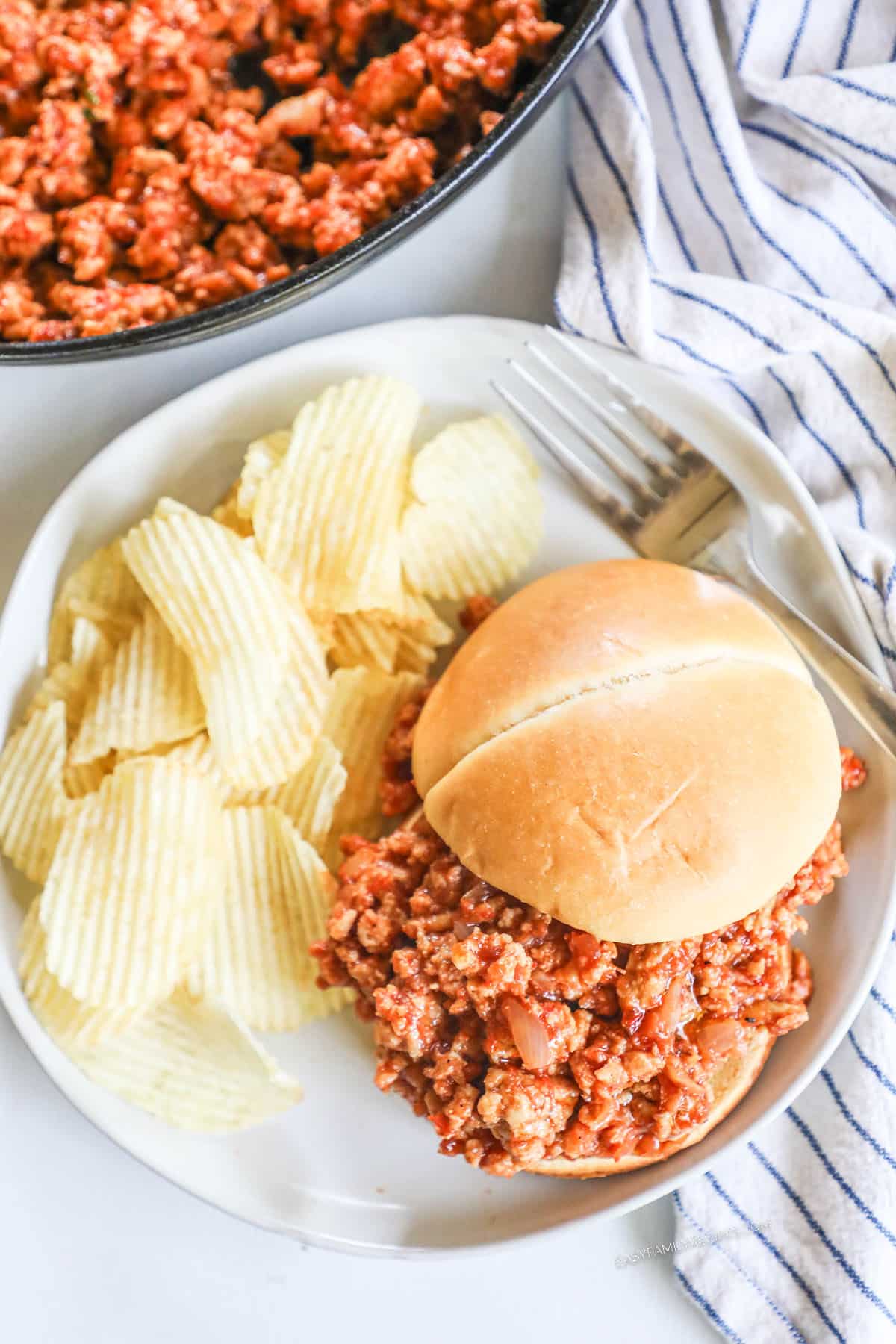 above image of a sloppy joe sandwich on a plate with a side of potato chips. 