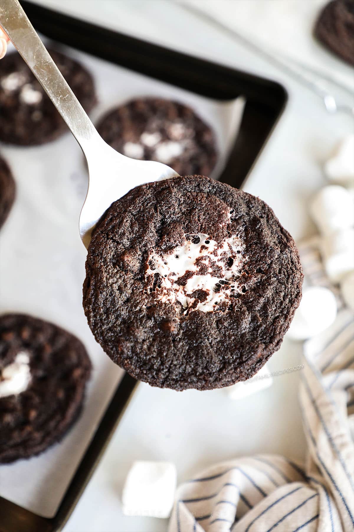 A silver spatula holds a Chocolate Marshmallow Cookie, with a baking sheet in the background holding more cookies.