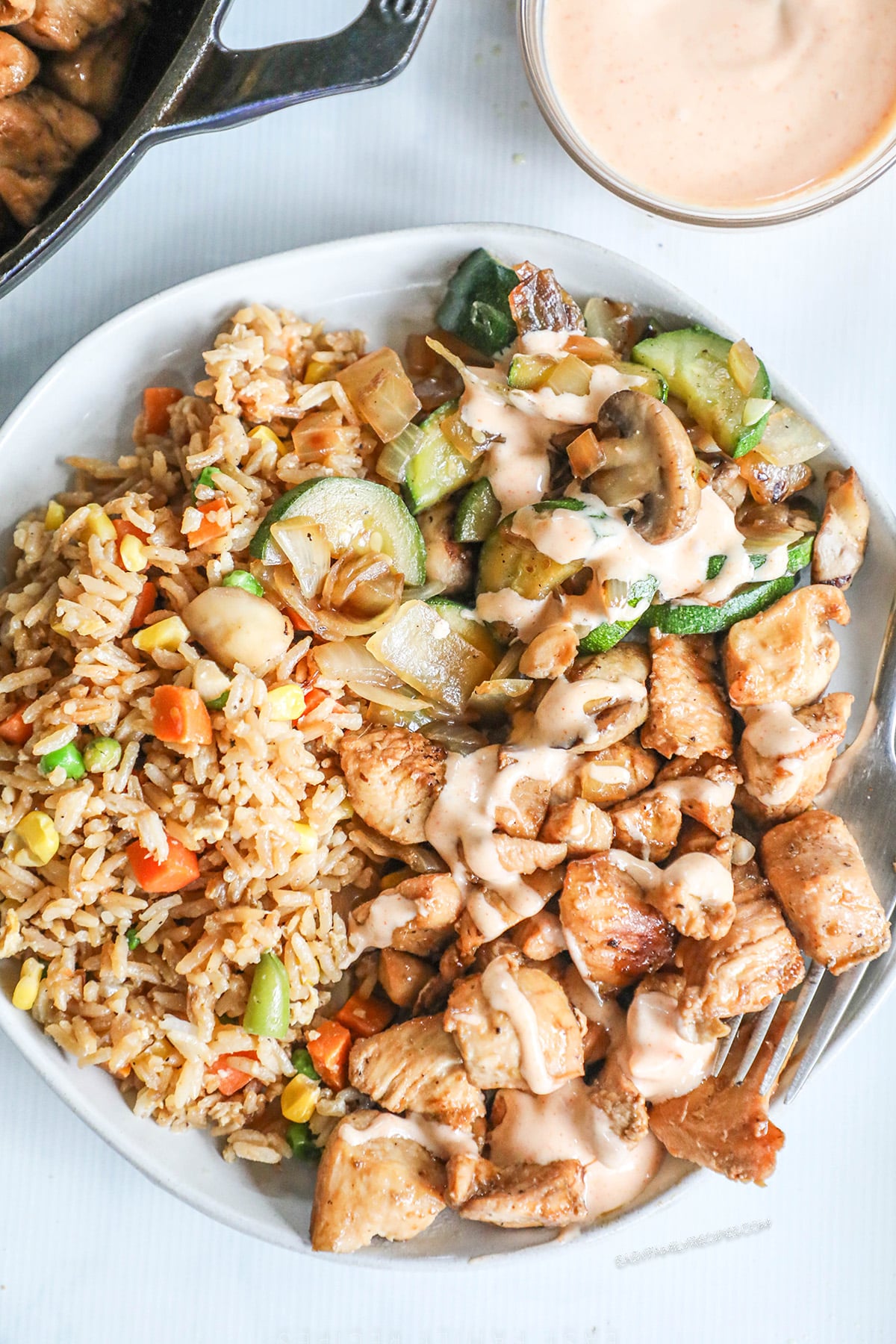 A white plate with Hibachi Chicken, vegetables, and rice along with sauce in the background.