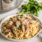 A serving of Crockpot Beef Stroganoff with noodles on a white plate with a silver fork. Parsley and a Crock Pot are in the background.