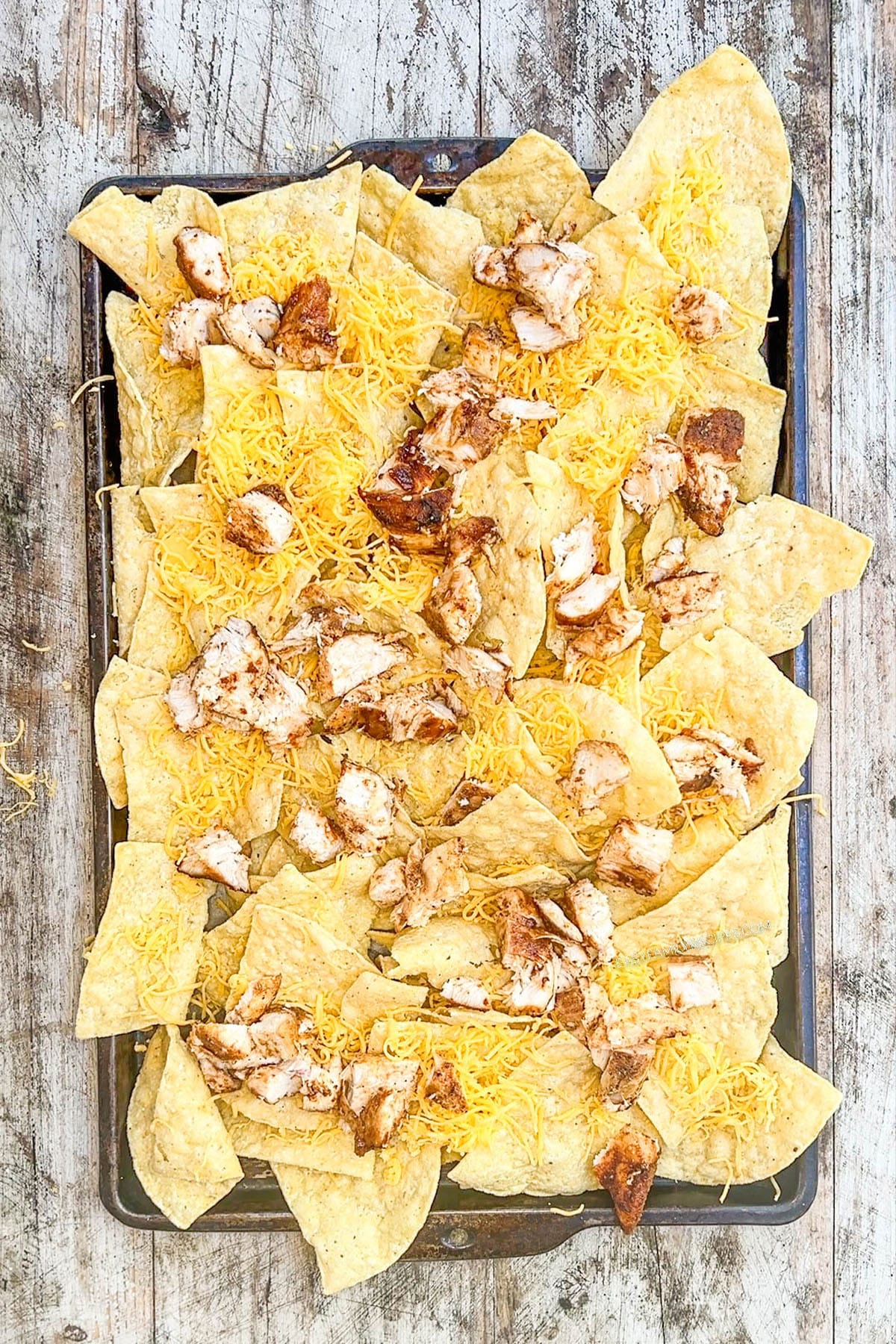 BBQ chicken is added to tortilla chips on a baking sheet, step 2 of making BBQ chicken nachos.