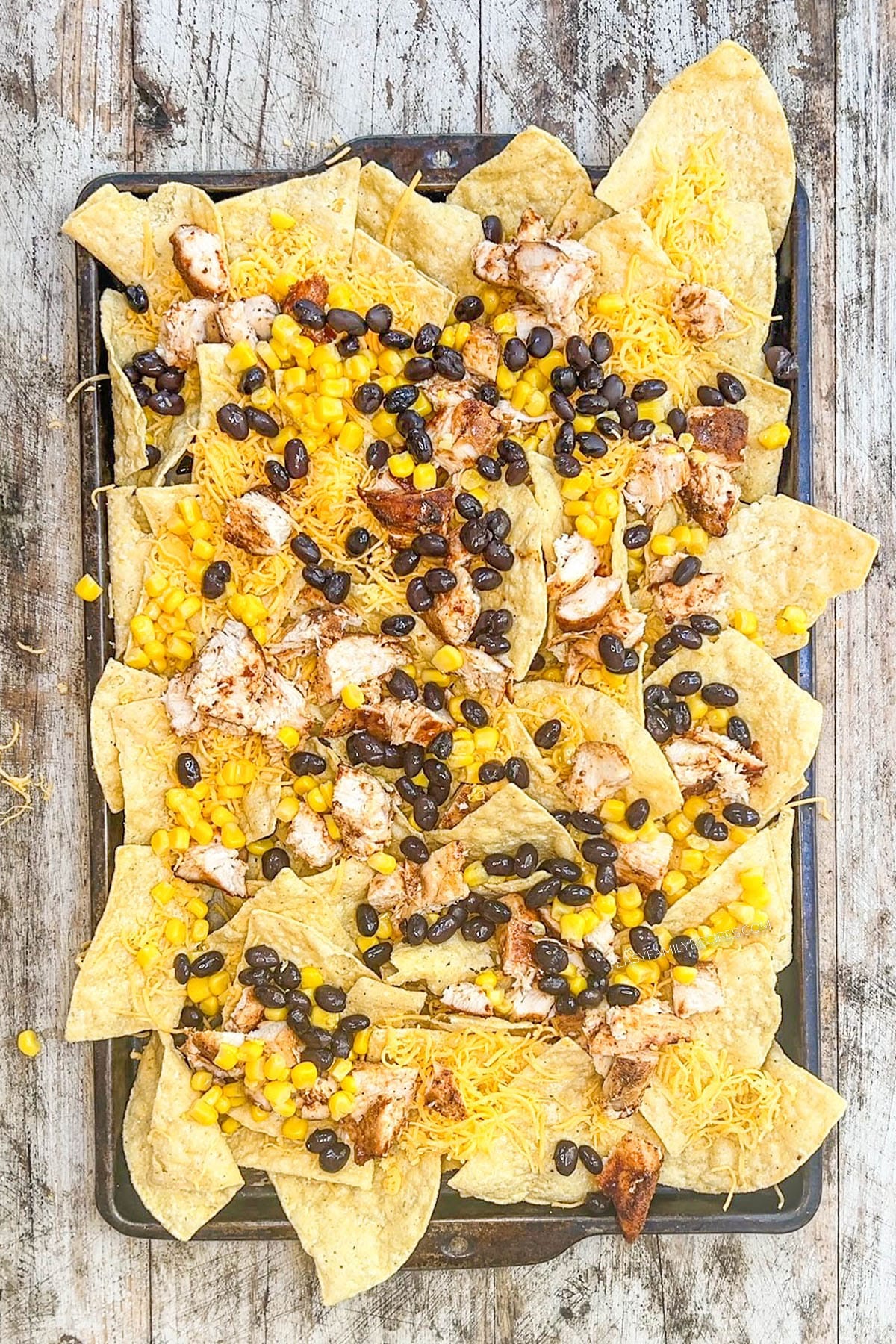 Toppings are added to tortilla chips on a baking sheet, step 3 of how to make BBQ chicken nachos.