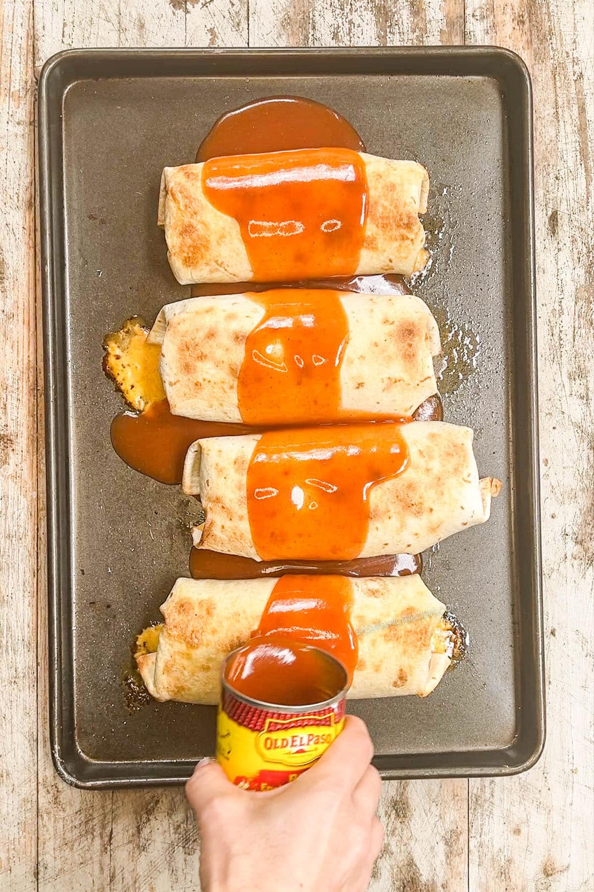 Step 5 of making chimichangas: Enchilada sauce is poured over baked chimichangas on a metal baking sheet.