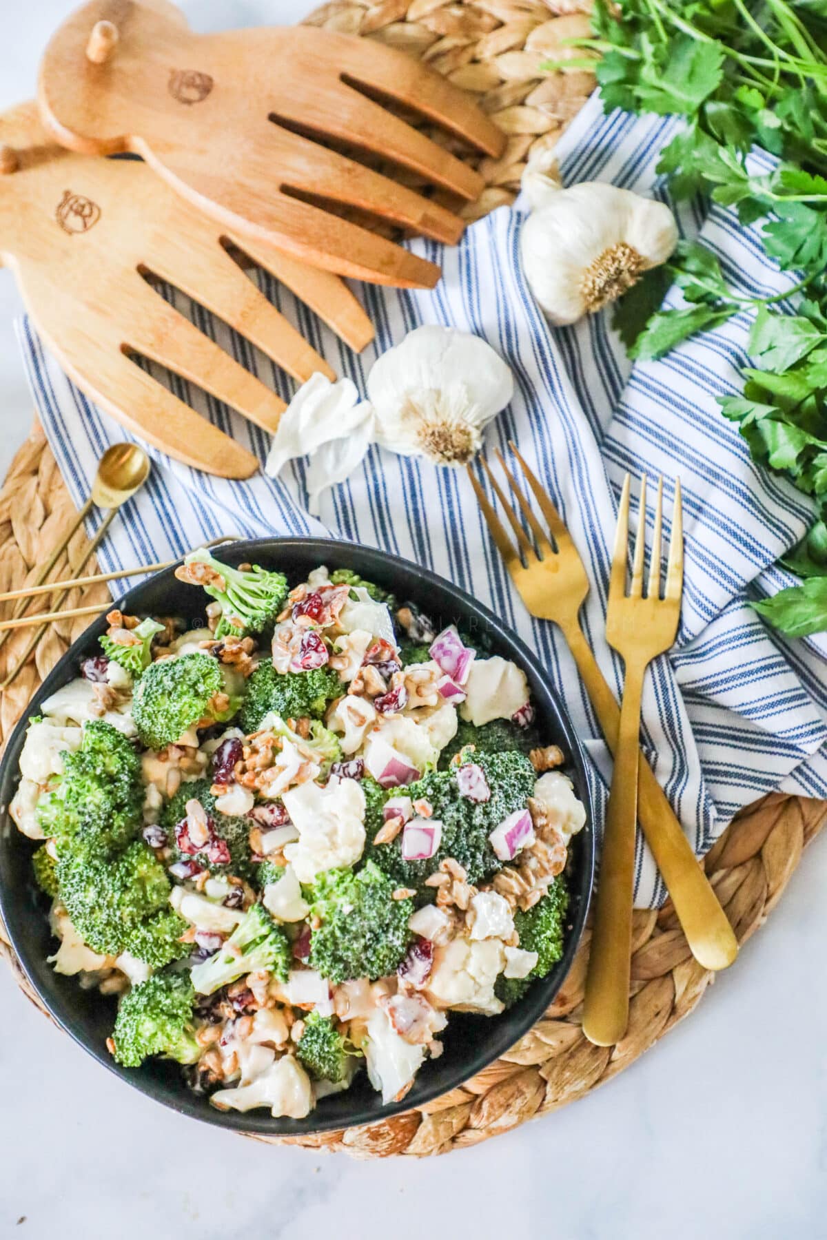 OVerhead shot of broccoli cauliflower salad. Salad tongs and gold utensils are next to the salad