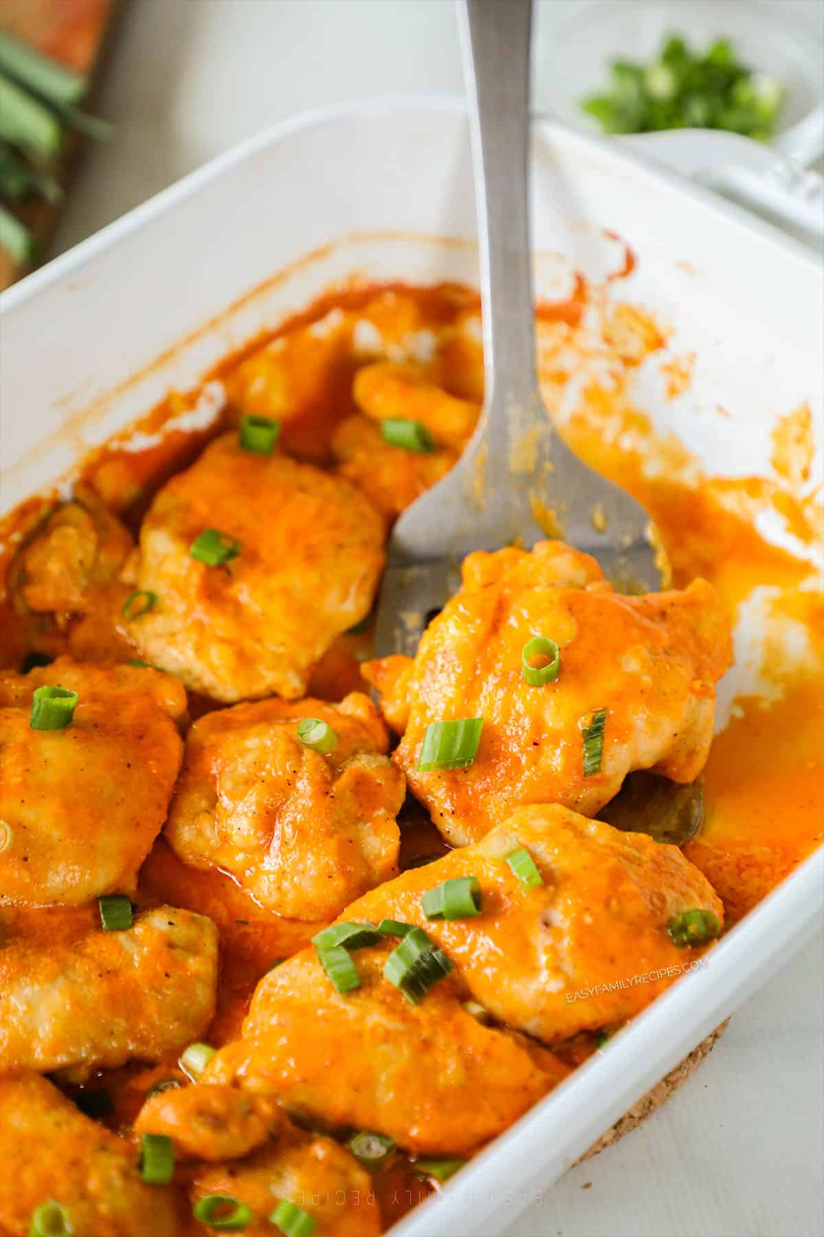 A white casserole dish full of Baked Buffalo Chicken Thighs being scooped out with a silver utensil.