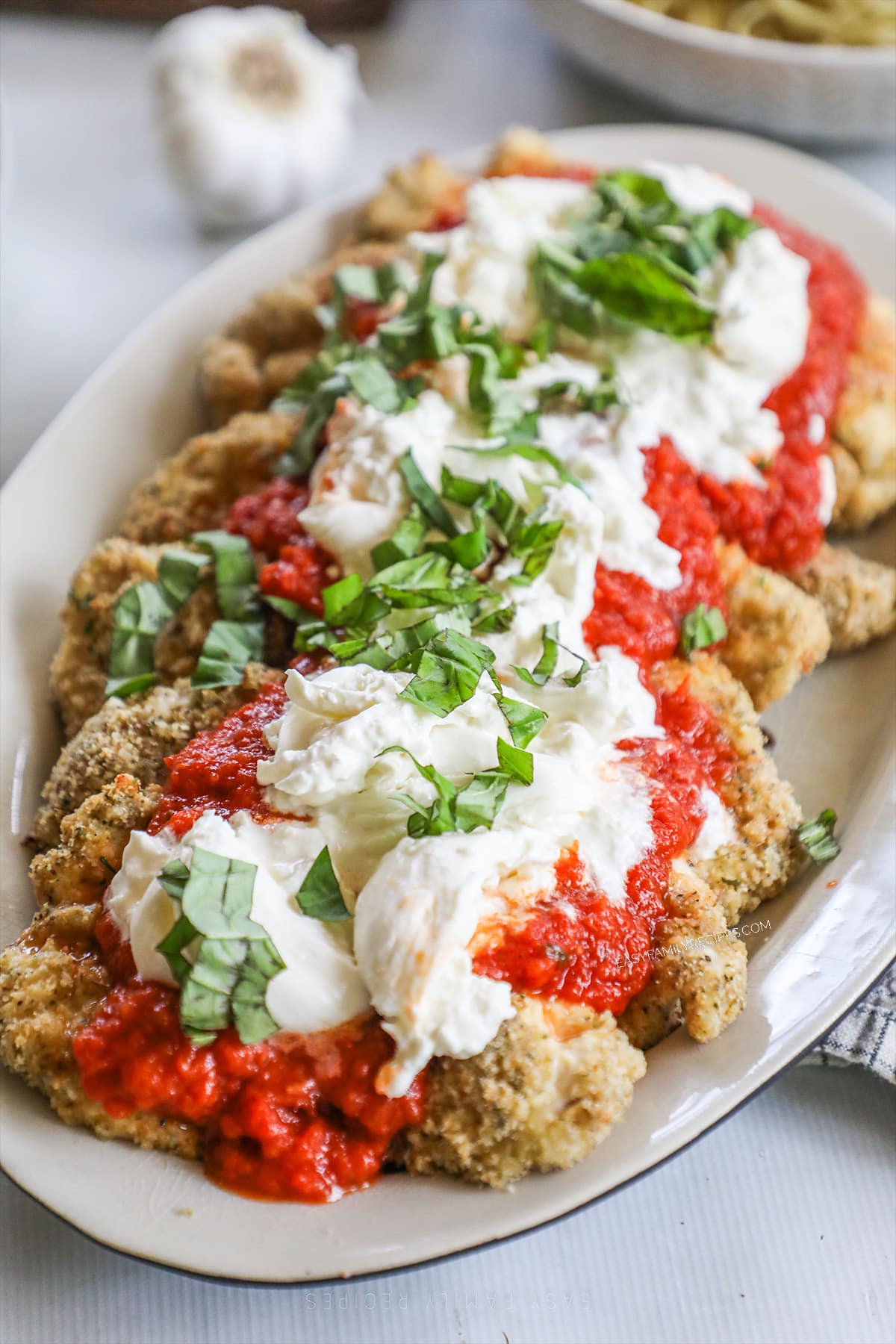 Burrata Chicken Parmesan on a white platter with garlic in the background. 