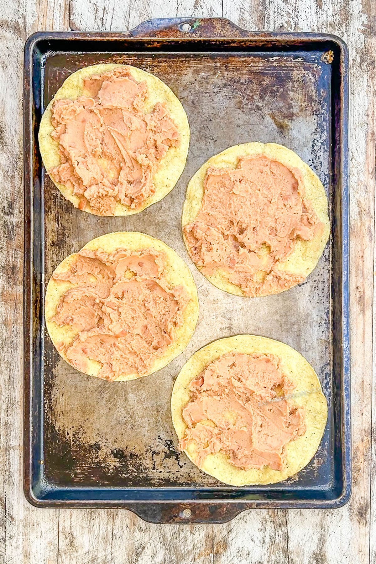 Top view of corn tortillas spread with refried beans on a baking sheet, for BBQ chicken tostadas.