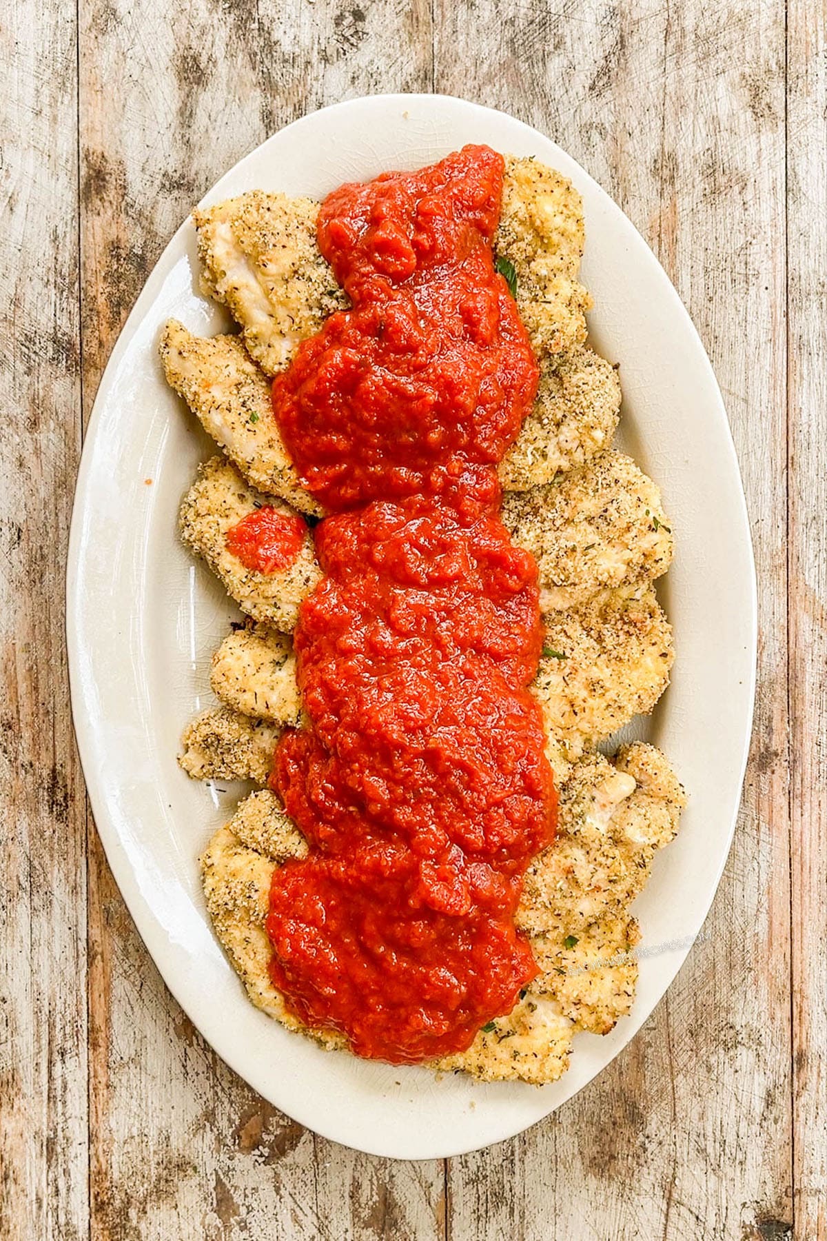 Marinara sauce is poured onto chicken cutlets on a white oval platter for Burrata Chicken Parmesan.