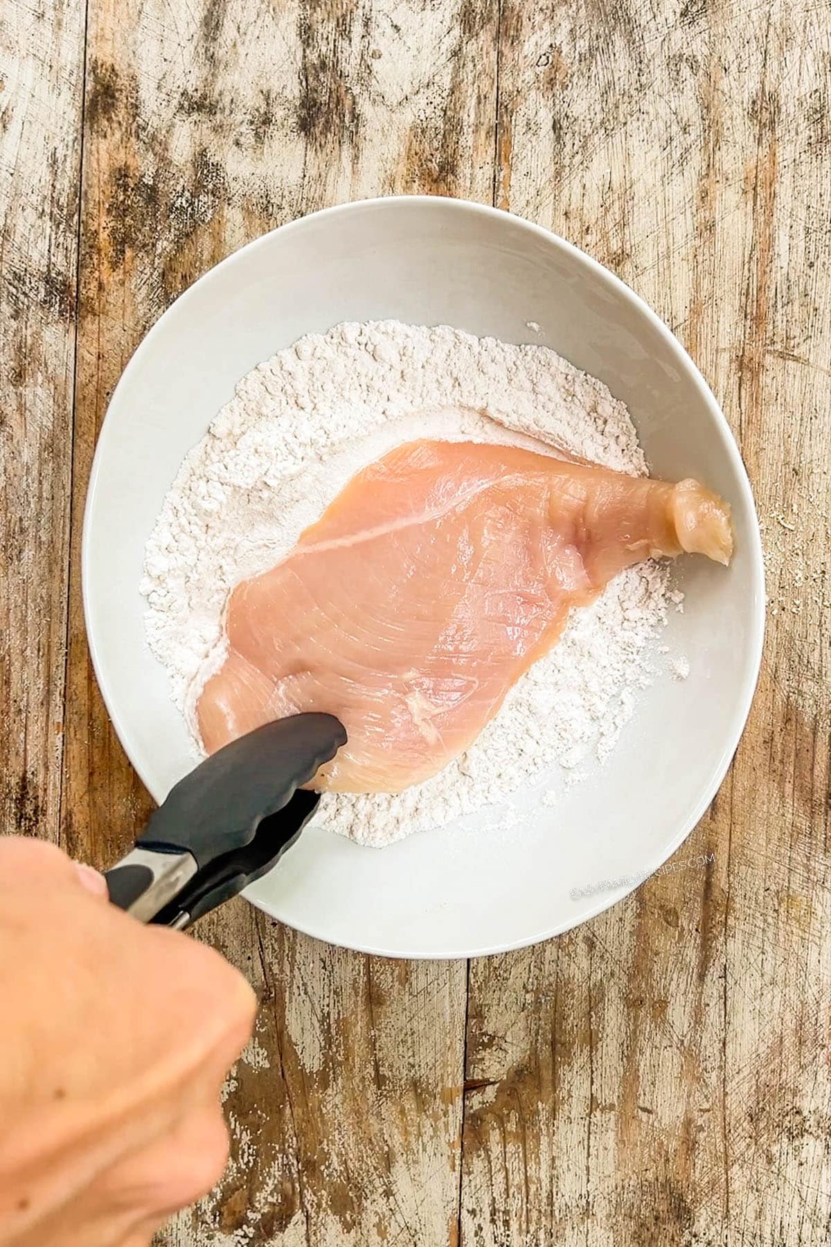 Chicken breast is dipped in flour in a white bowl for baked chicken cutlets.