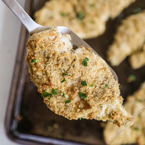 A silver serving spoon scoops a baked chicken cutlet off of a baking sheet.