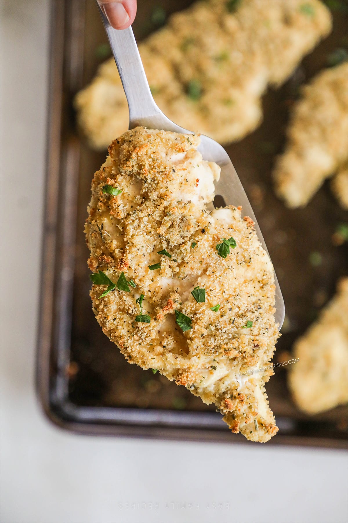 A silver serving spoon scoops a baked chicken cutlet off of a baking sheet.