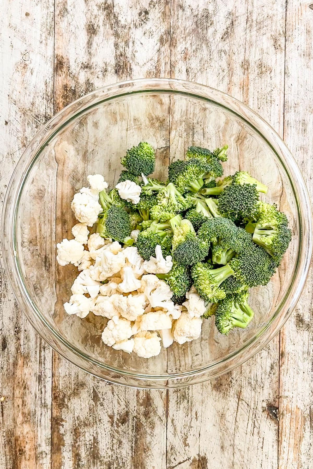 Broccoli and cauliflower are added to a clear glass bowl for broccoli crunch salad.