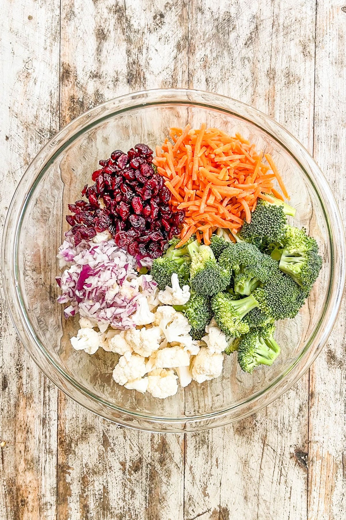 Carrots, broccoli, cauliflower, red onion, and dried cranberries in a bowl for A white bowl filled with Broccoli Crunch Salad.