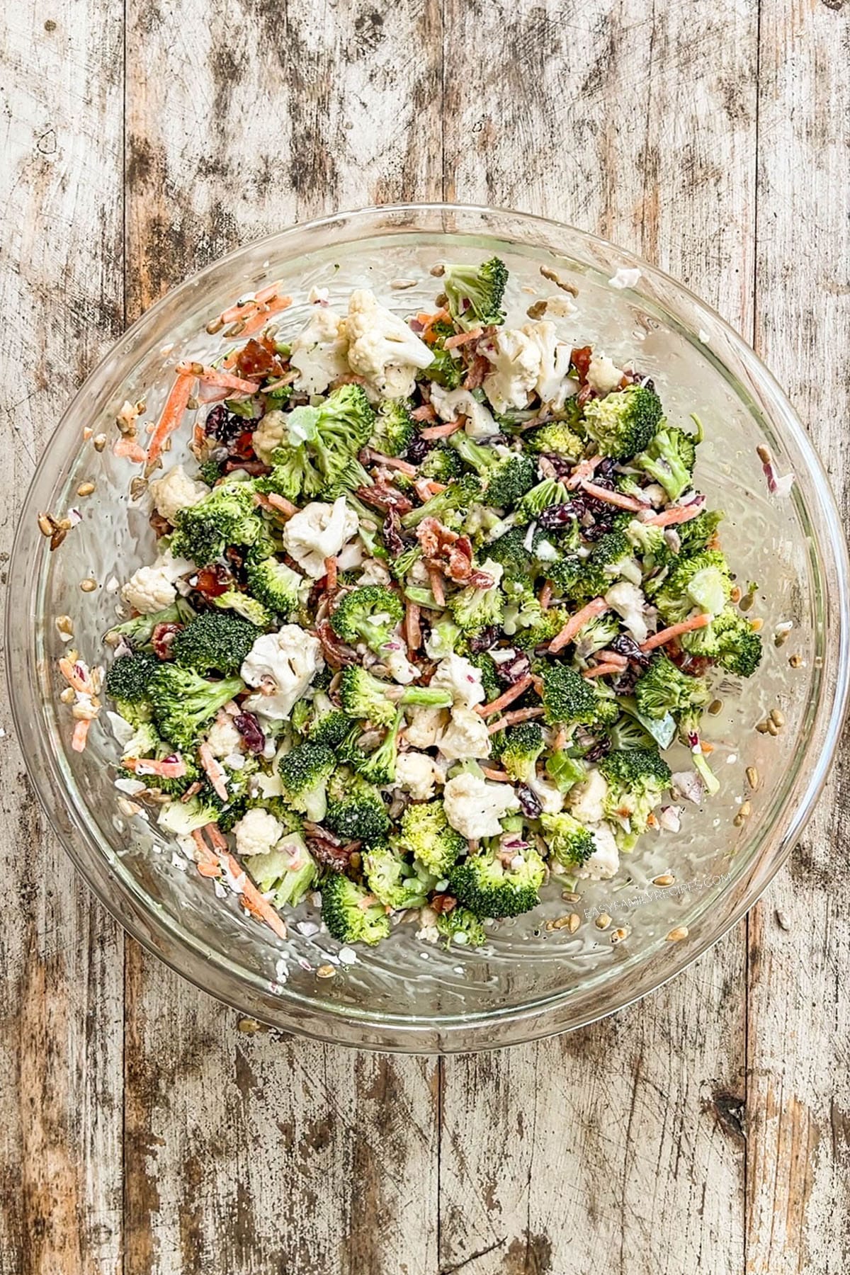 Prepared A white bowl filled with Broccoli Crunch Salad in a clear glass mixing bowl on a distressed wood background.