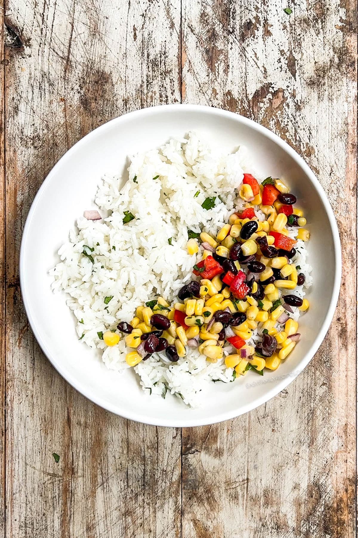 Top view of a white bowl with cilantro lime rice and black bean and corn salsa, forming the base of a Southwest Chicken Bowl. The ingredients are in a white ceramic dish on a distressed wood background.