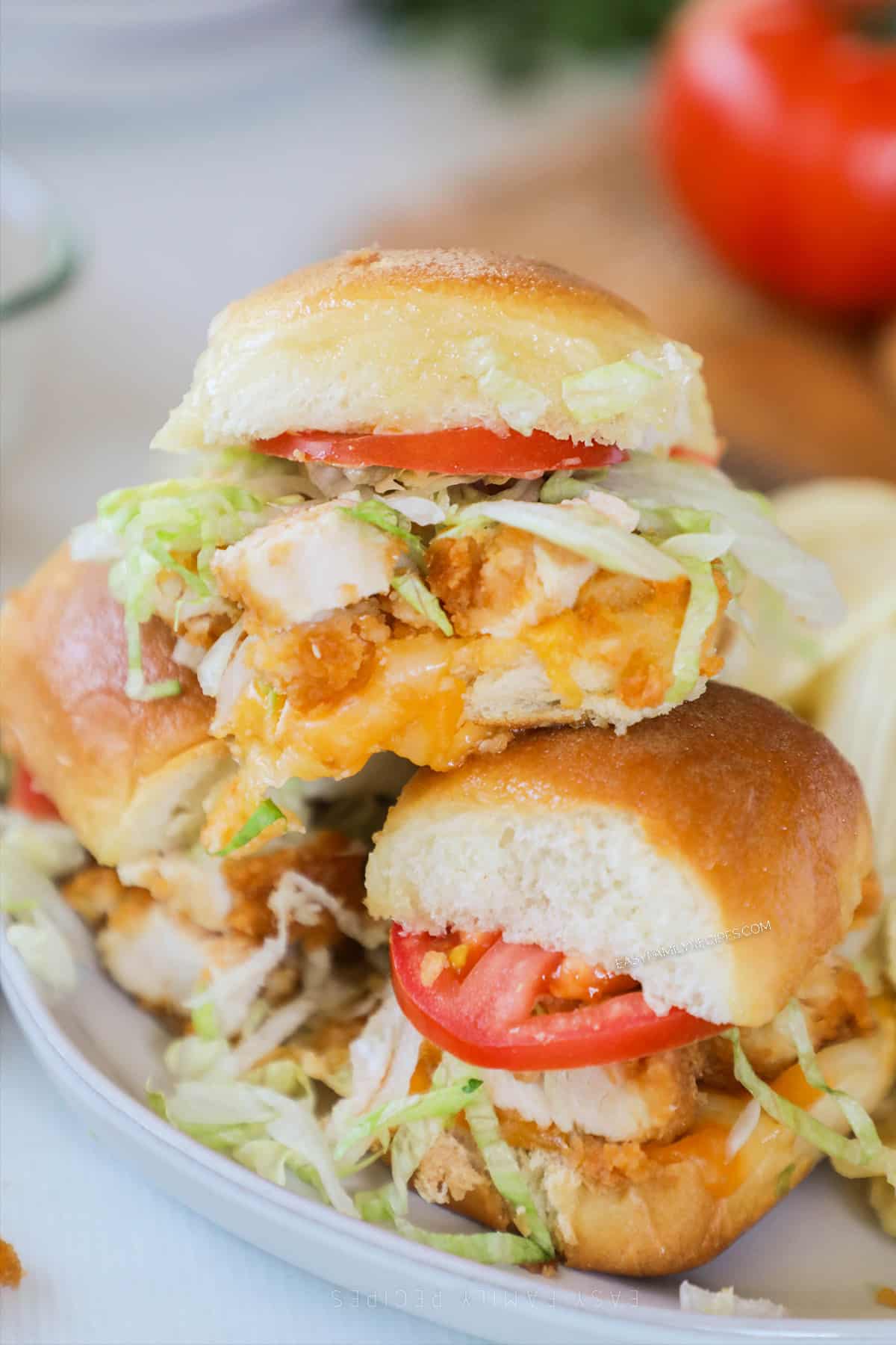 A pile of chicken tender sliders on a white plate with potato chips and a tomato in the background.