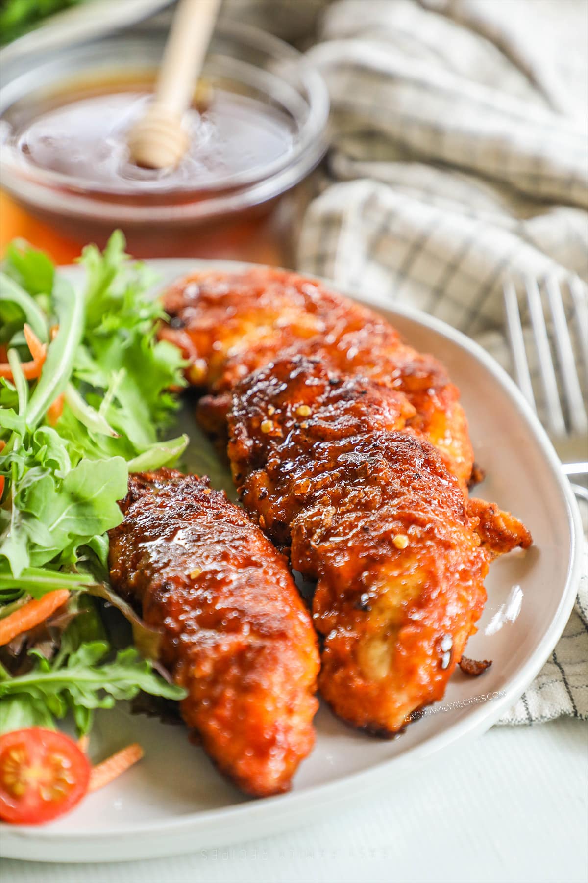 A white platter full of hot honey chicken tenders with fresh herbs and a salad