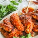 A white platter full of hot honey chicken tenders with fresh herbs. A glass cup full of hot honey with a honey dipper is in the background. The text reads, “Hot Honey Chicken Tenders”