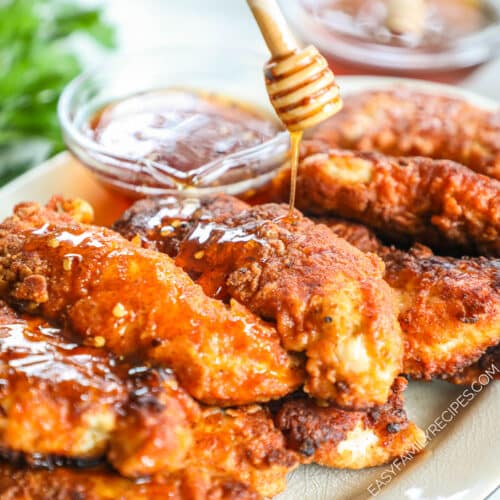 A white platter full of hot honey chicken tenders with fresh herbs. A honey dipper drizzles hot honey over the freshly fried chicken.