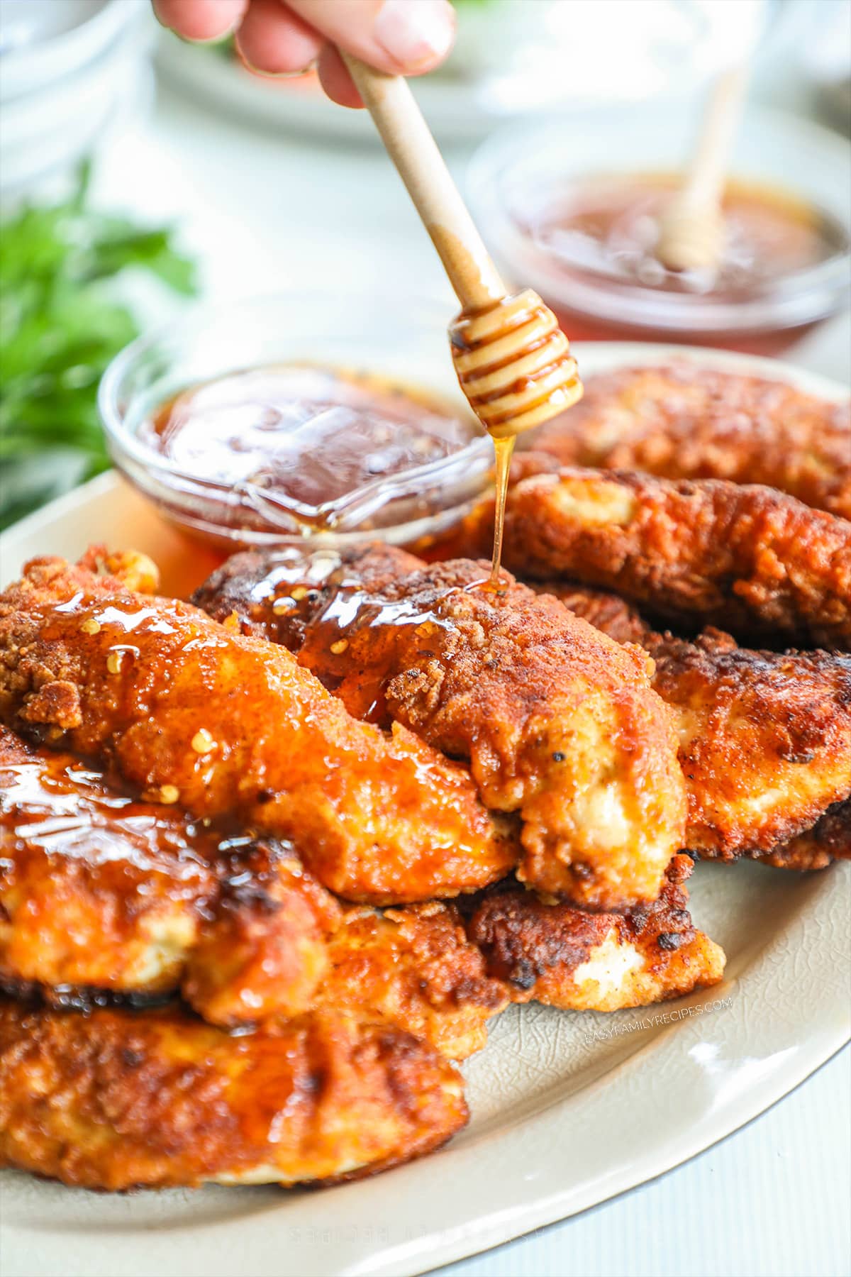 A white platter full of hot honey chicken tenders with fresh herbs. A honey dipper drizzles hot honey over the freshly fried chicken. 