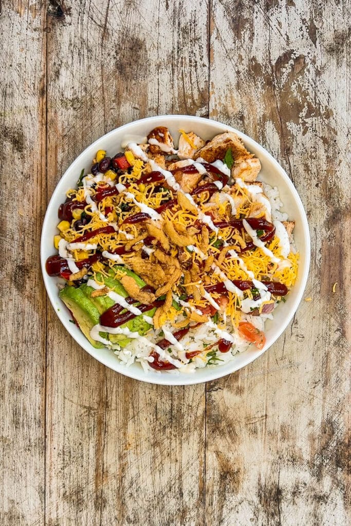 French fried onions and ranch dressing are added to a BBQ chicken bowl in a white bowl on a distressed wood background.