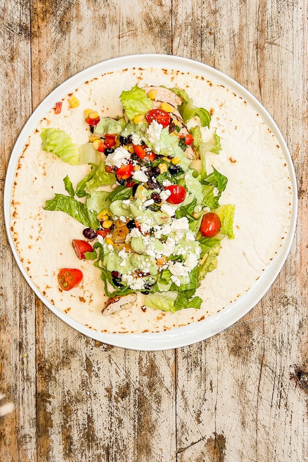 a tortilla on a plate topped with baja chicken, lettuce, tomato, and dressing.