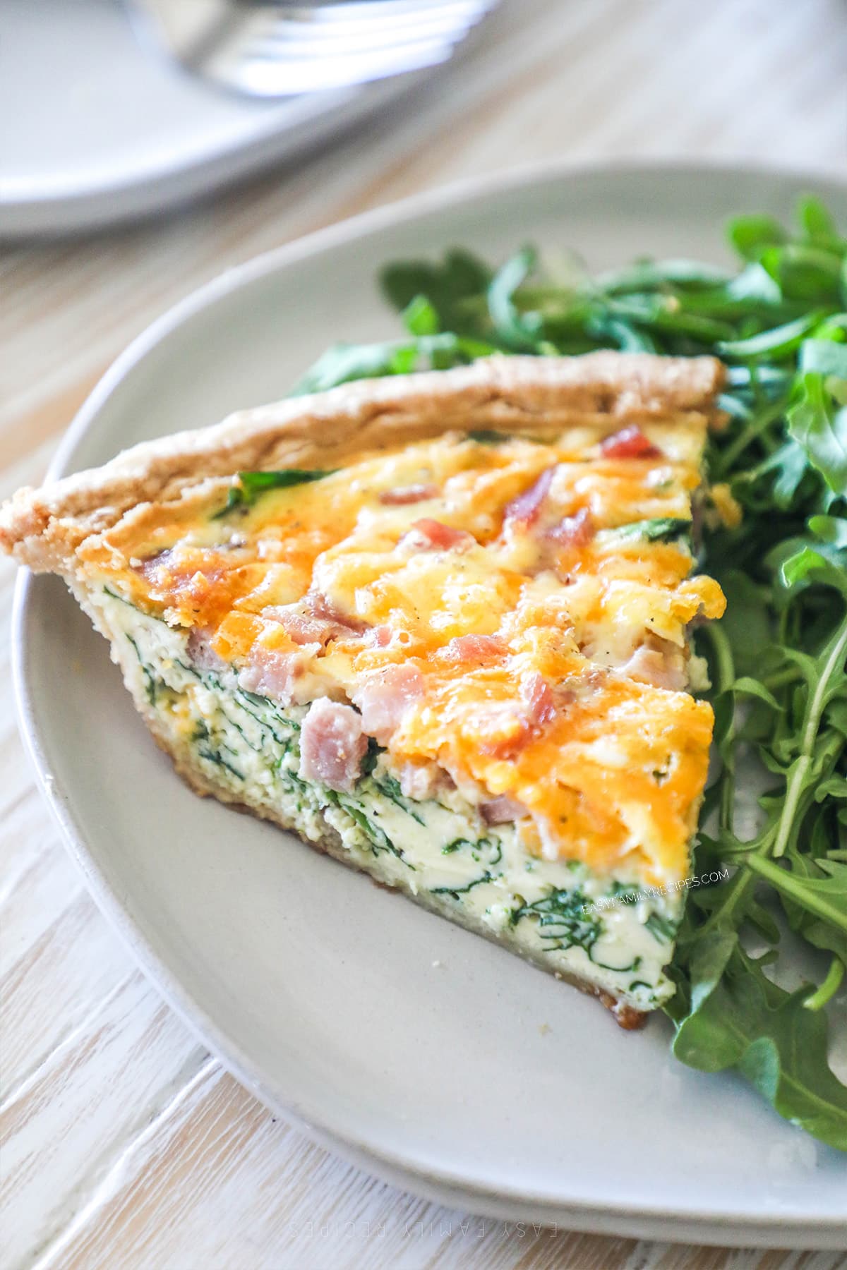 A slice of ham and spinach quiche on a white plate with a salad alongside.