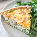 A slice of ham and spinach quiche on a white plate with a salad alongside.