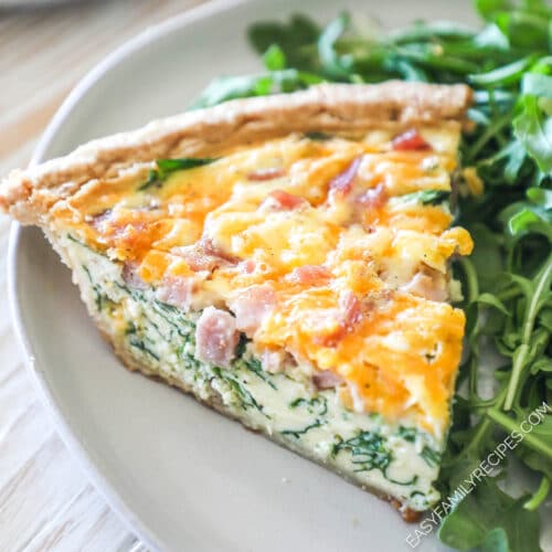 A slice of ham and spinach quiche on a white plate with a salad alongside.