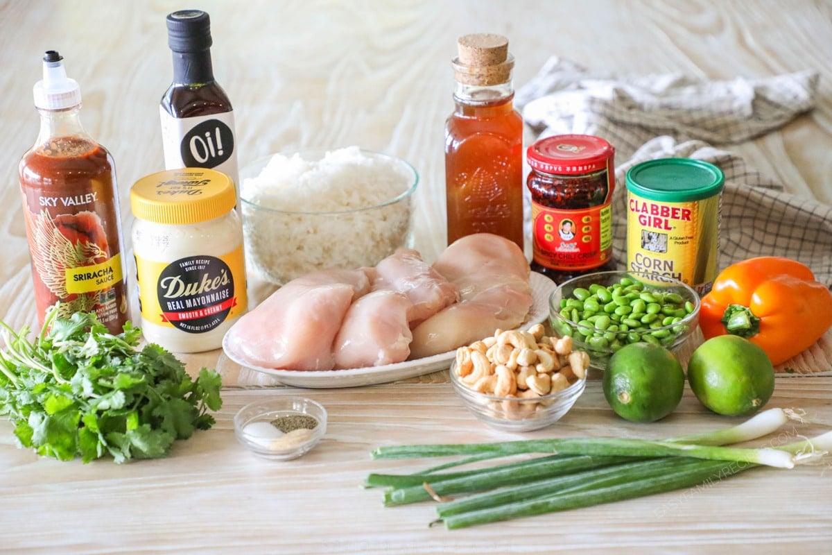 Ingredients for recipe before prepping: sriracha sauce, mayo, fresh cilantro, oil, cooked rice, raw chicken breasts, honey, chili crisp, corn starch, cashews, edamame, lime, bell pepper, seasonings, and green onion.