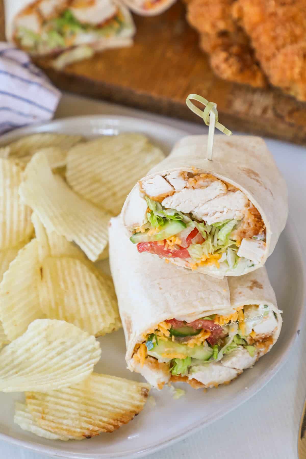A chicken tender wrap cut in half with a skewer tucked into the top. It's on a white plate with potato chips on the side.