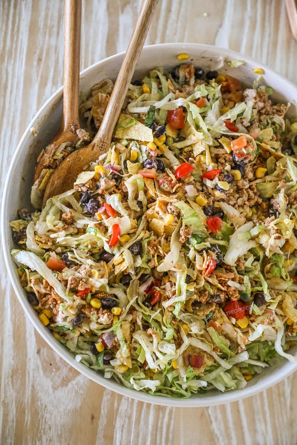 Top view of a white bowl full of layered taco salad with wooden salad spoons.