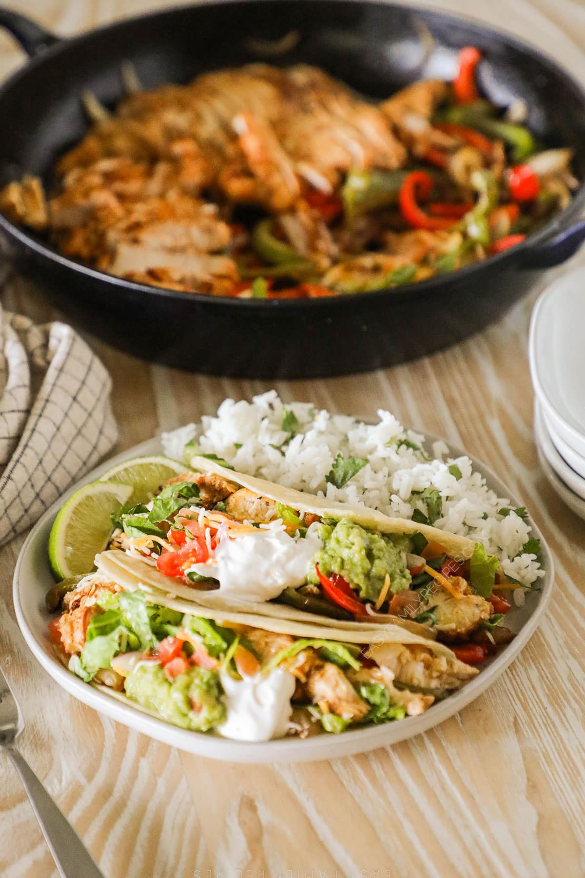 A white plate with Fajita Tacos and rice on a whitewashed wooden background. A skillet full of fajita chicken and veggies is in the background. 