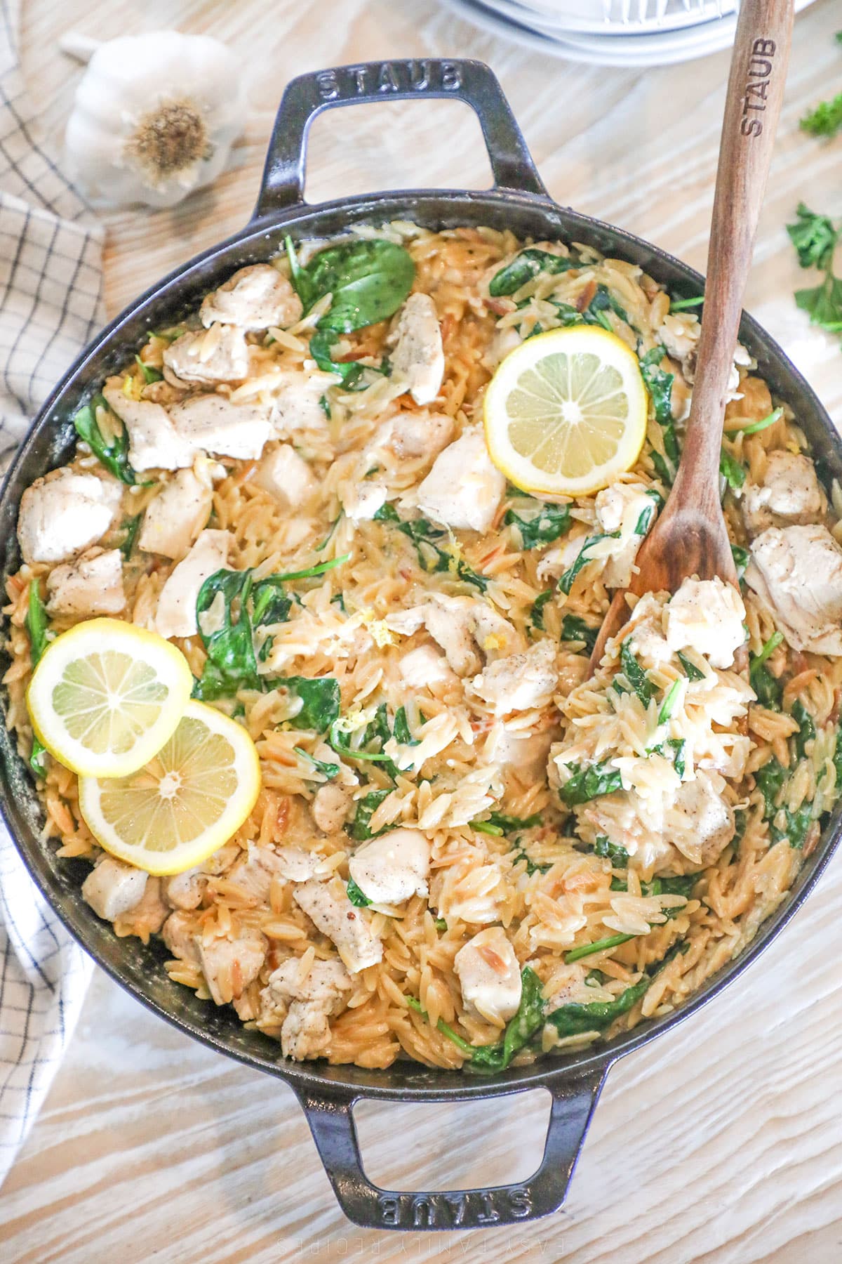Top view of creamy lemon chicken orzo in a skillet with a wooden spoon. Slices of lemon and herbs garnish the top. 
