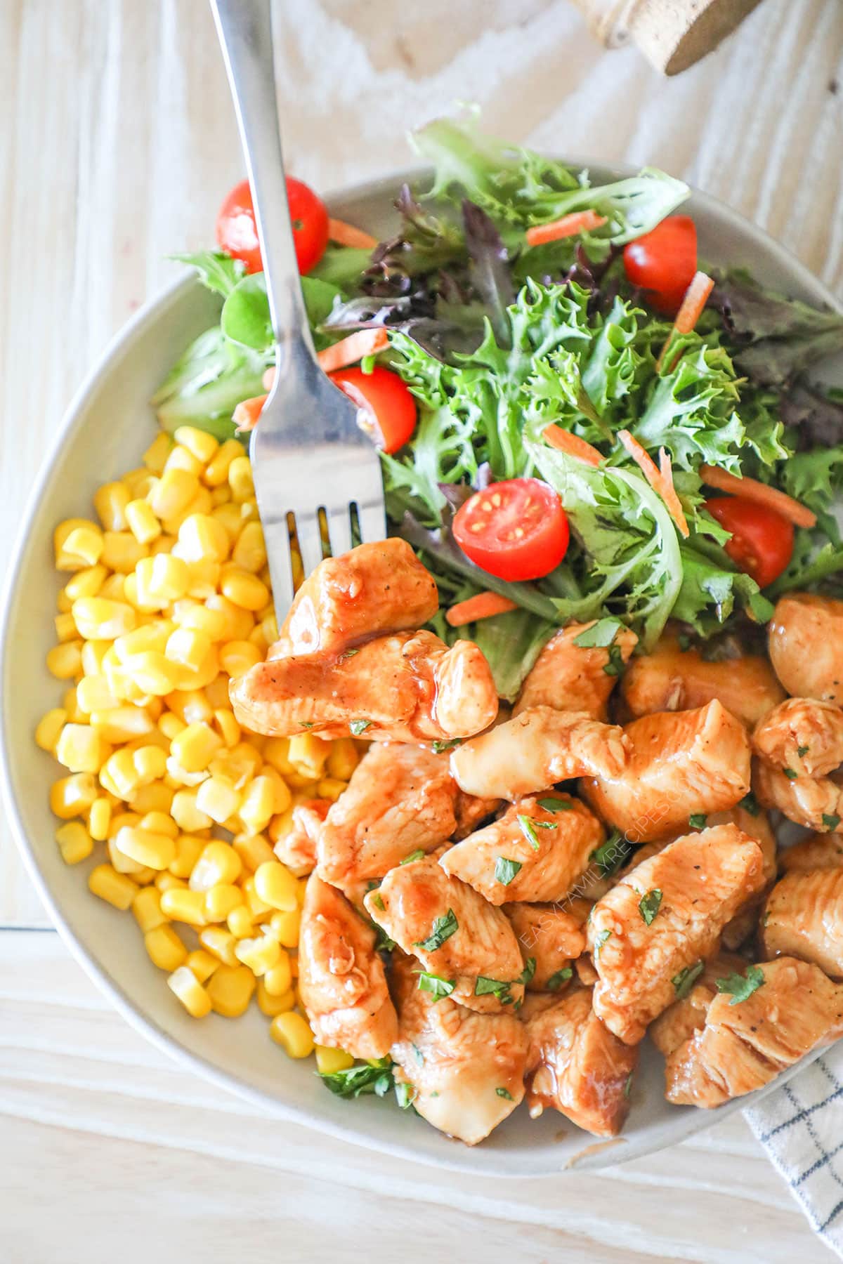 Top view of a white plate full of honey BBQ chicken bites, corn, and a simple green salad with a silver fork. 