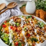 A white bowl full of chicken tender salad with a drizzle of ranch dressing and a bottle of dressing in the background. The text reads, “Chicken Tender Salad”