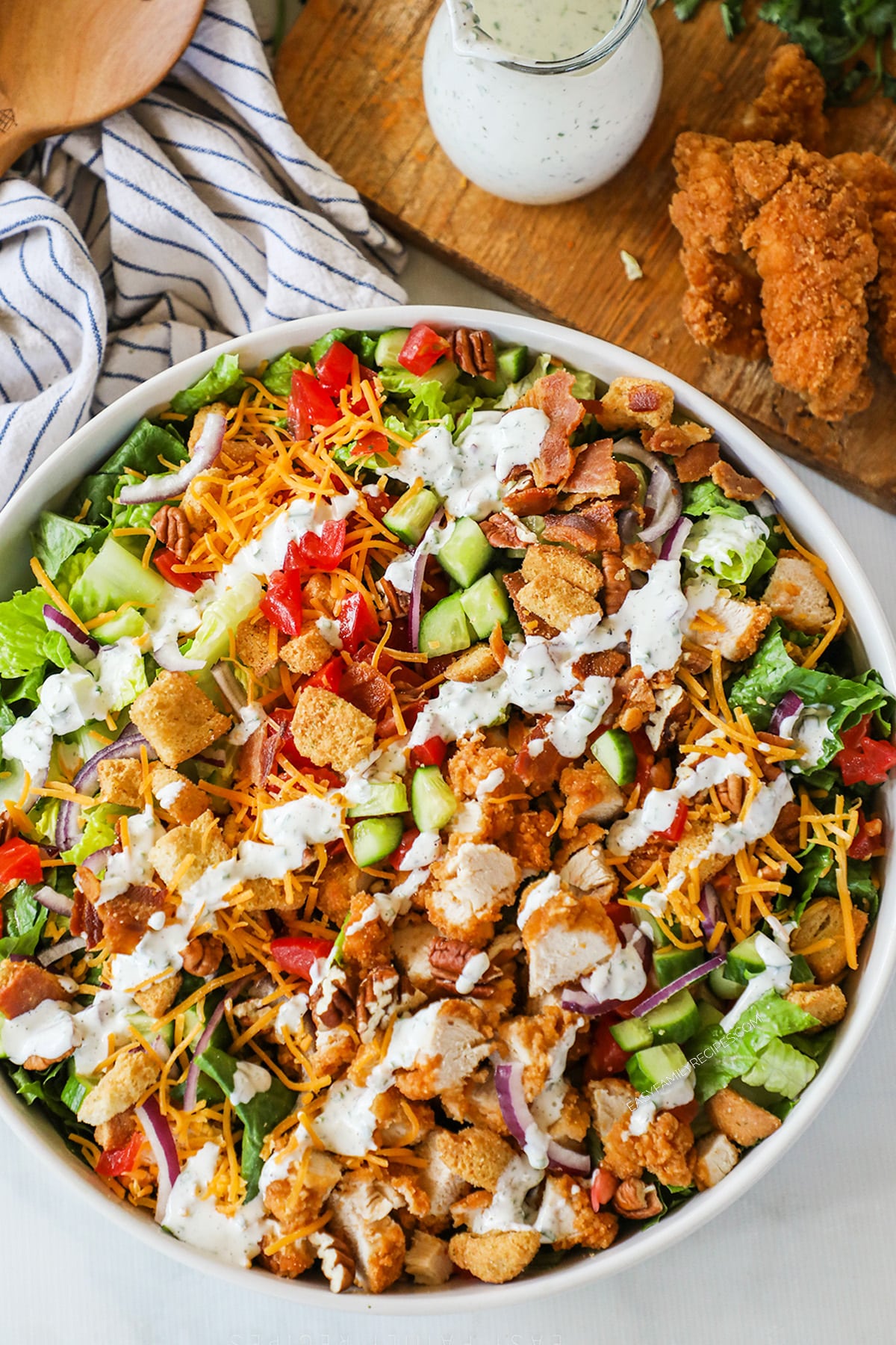 Top view of chicken tender salad in a white bowl with a drizzle of ranch dressing on a striped linen. 