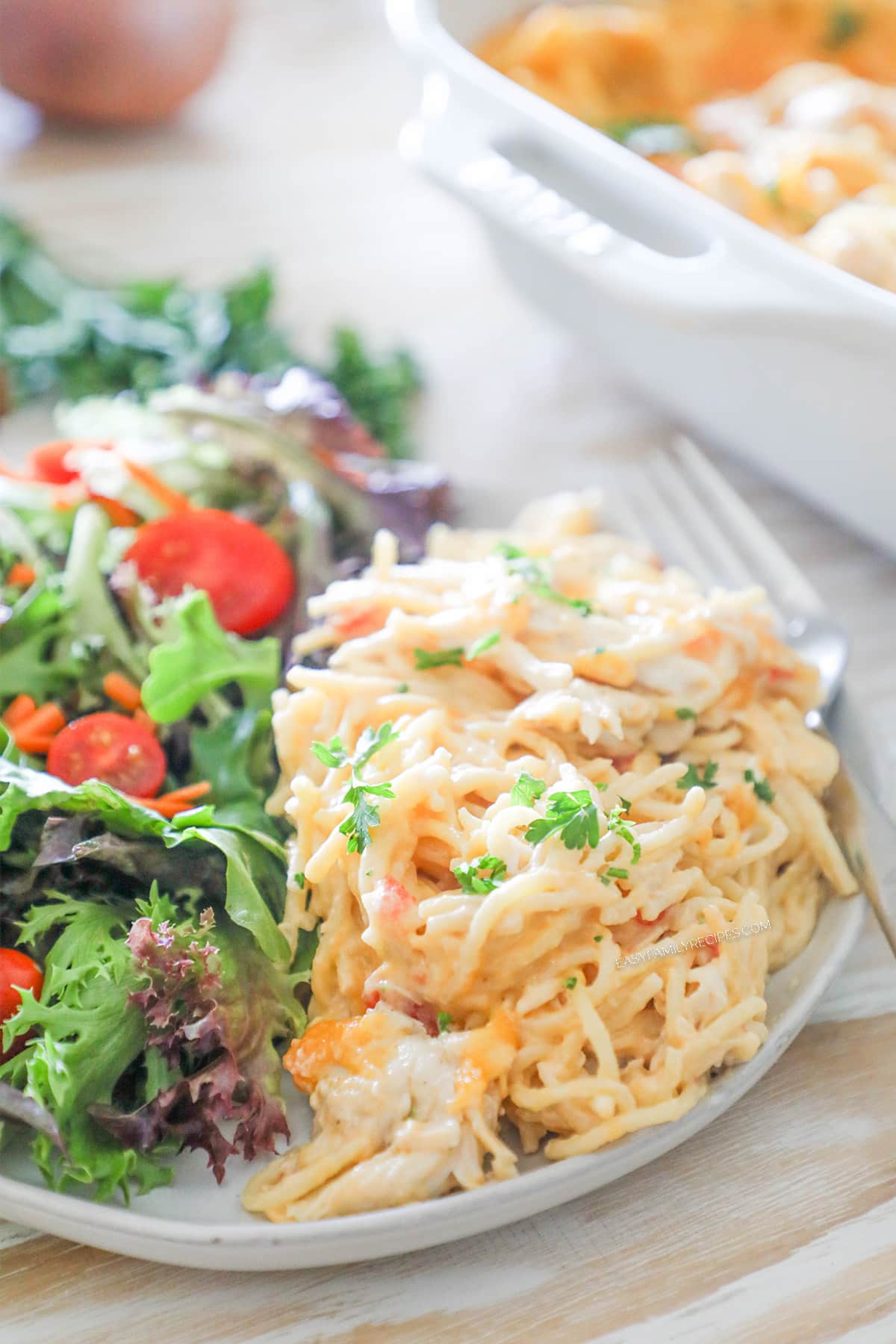A white plate with a serving of baked chicken spaghetti and a side salad on a whitewashed background.