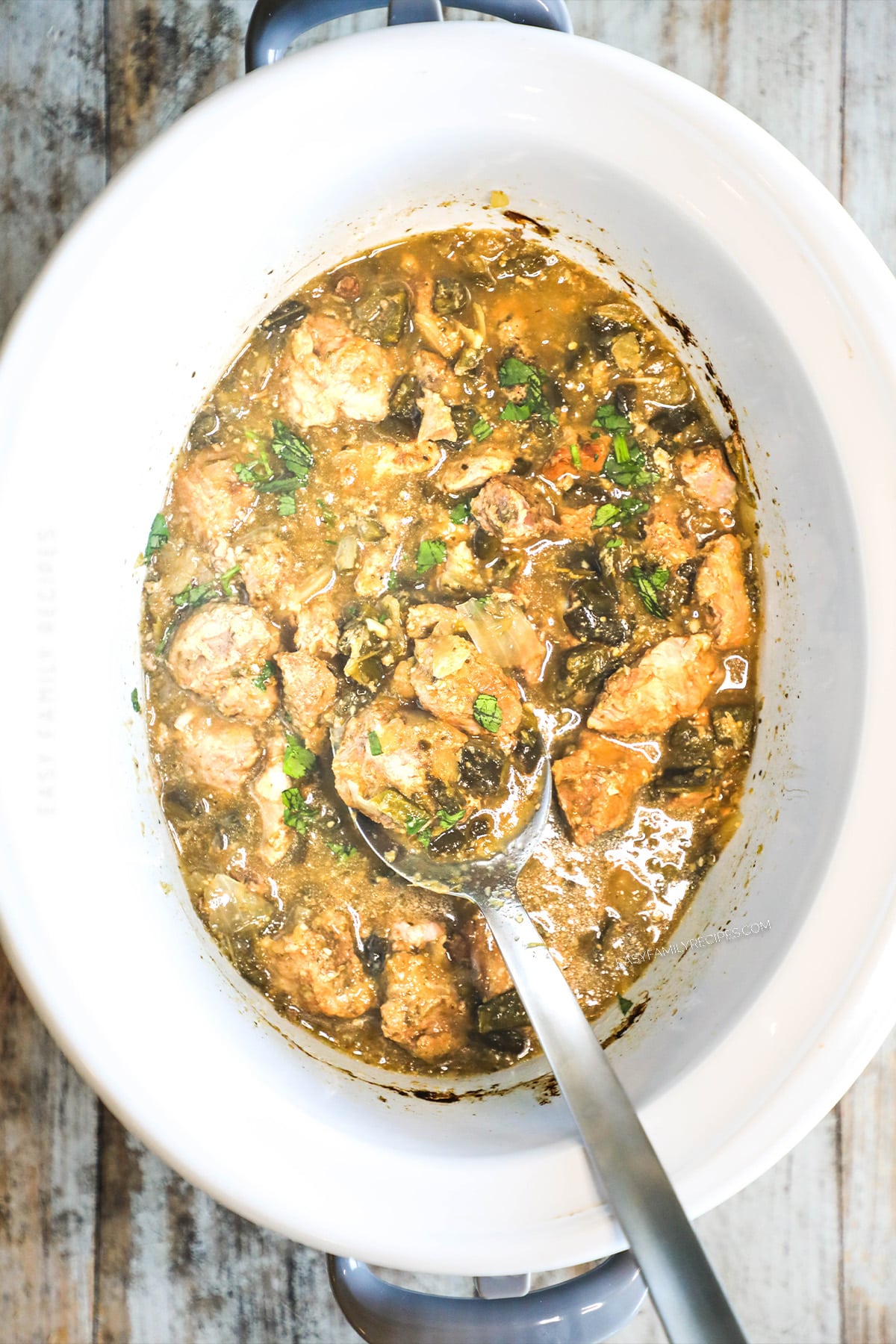 A top view of Crockpot Pork Chile Verde in a white slow cooker crock. A silver spoon is sitting in the chile verde.