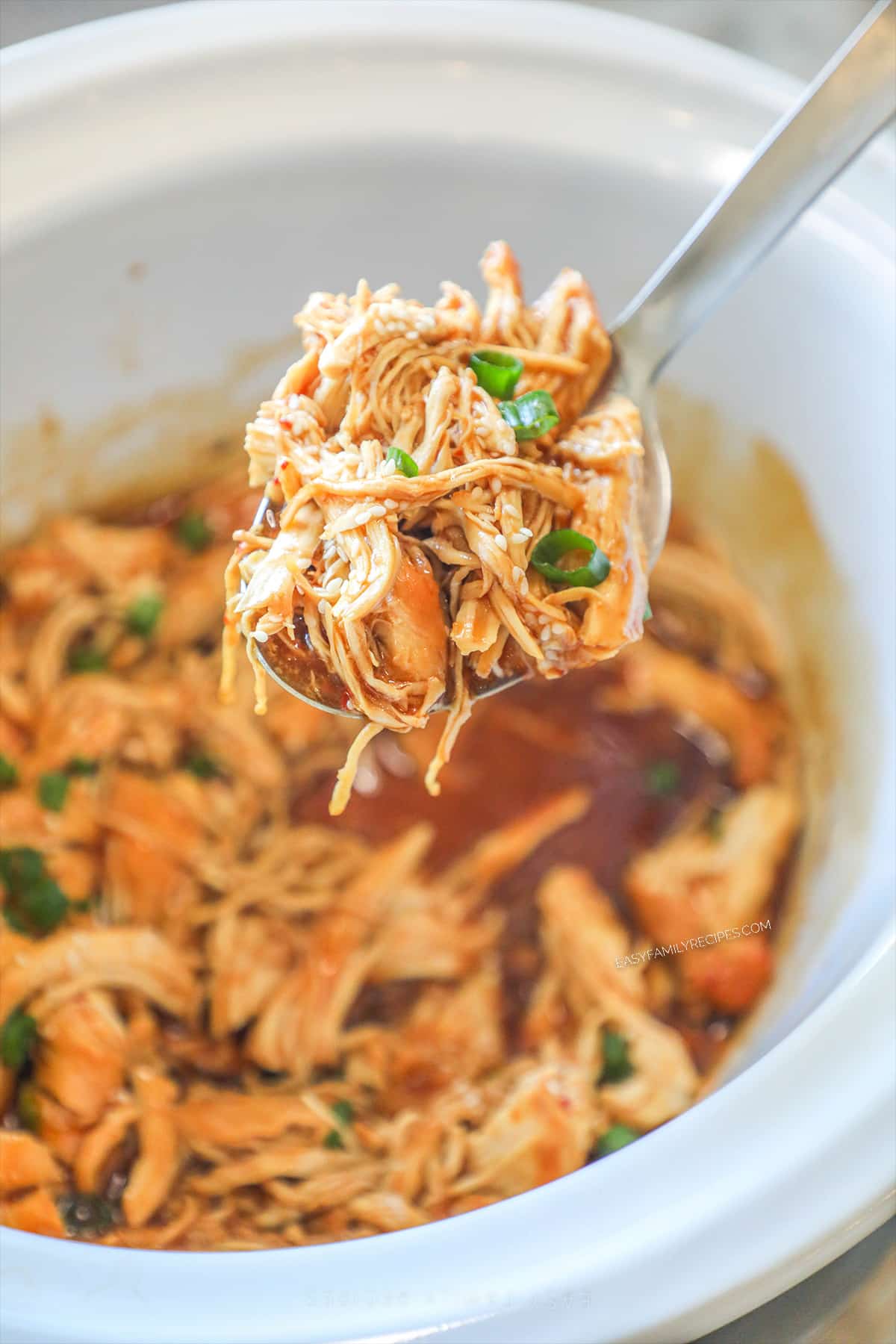 A spoon lifts Crockpot Bourbon chicken out of a white slow cooker. 