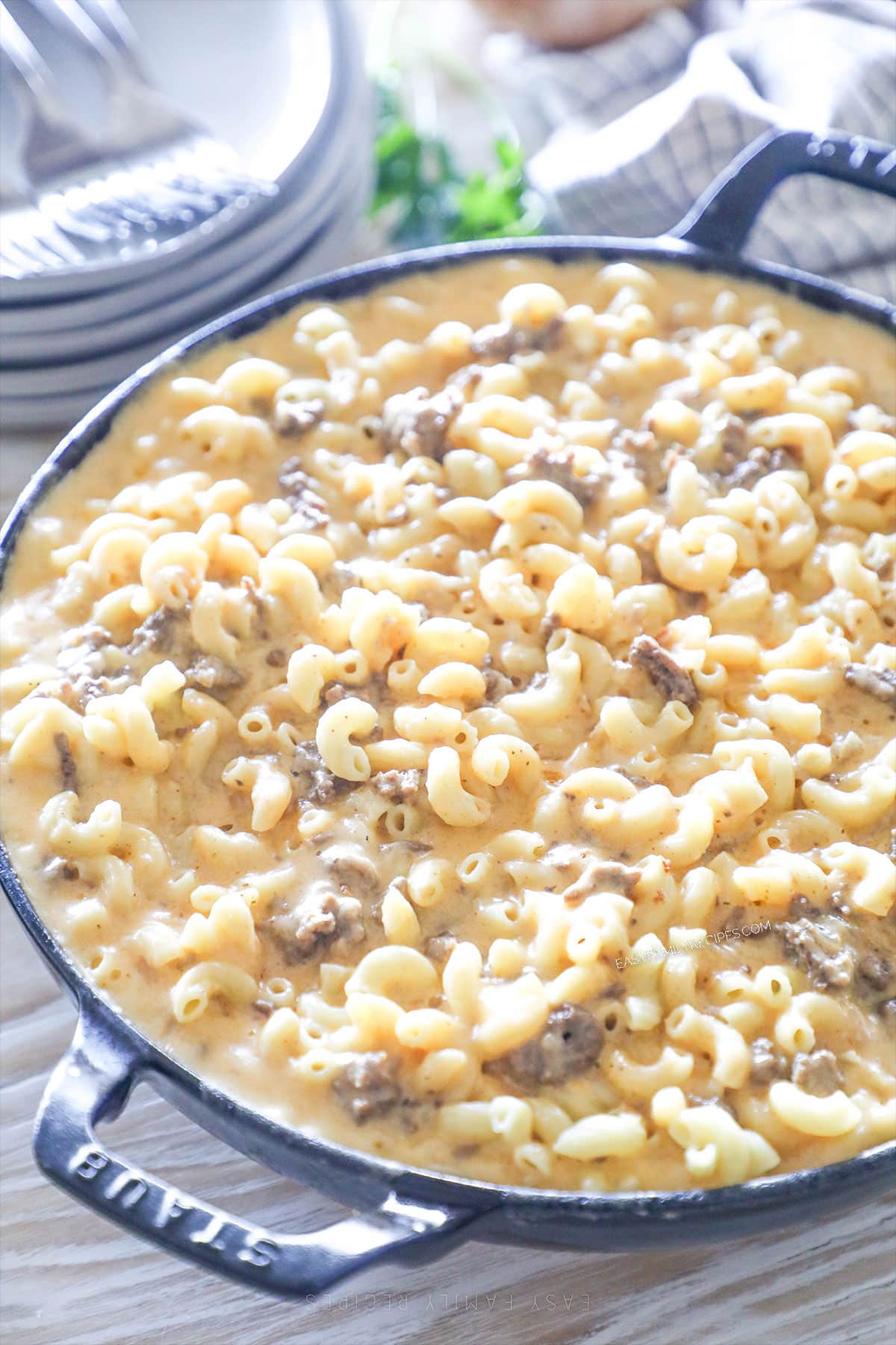 Loaded Mac & Cheese with Ground Beef in a blue skillet with a pile of plates in the background.