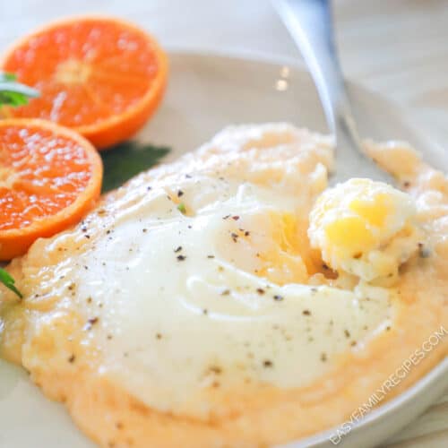 A white plate with cheesy grits and eggs along with an orange and parsley for garnish.