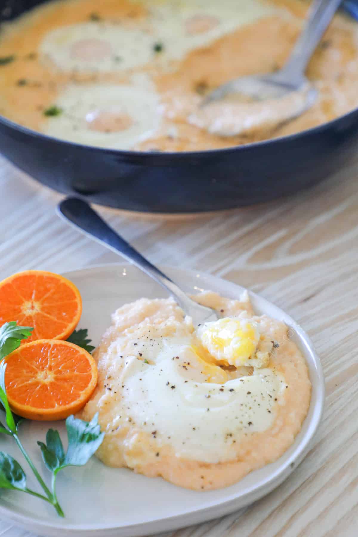 A white plate with cheesy grits and eggs along with an orange and parsley for garnish. A cast-iron pan full of cheesy grits with eggs are in the background.