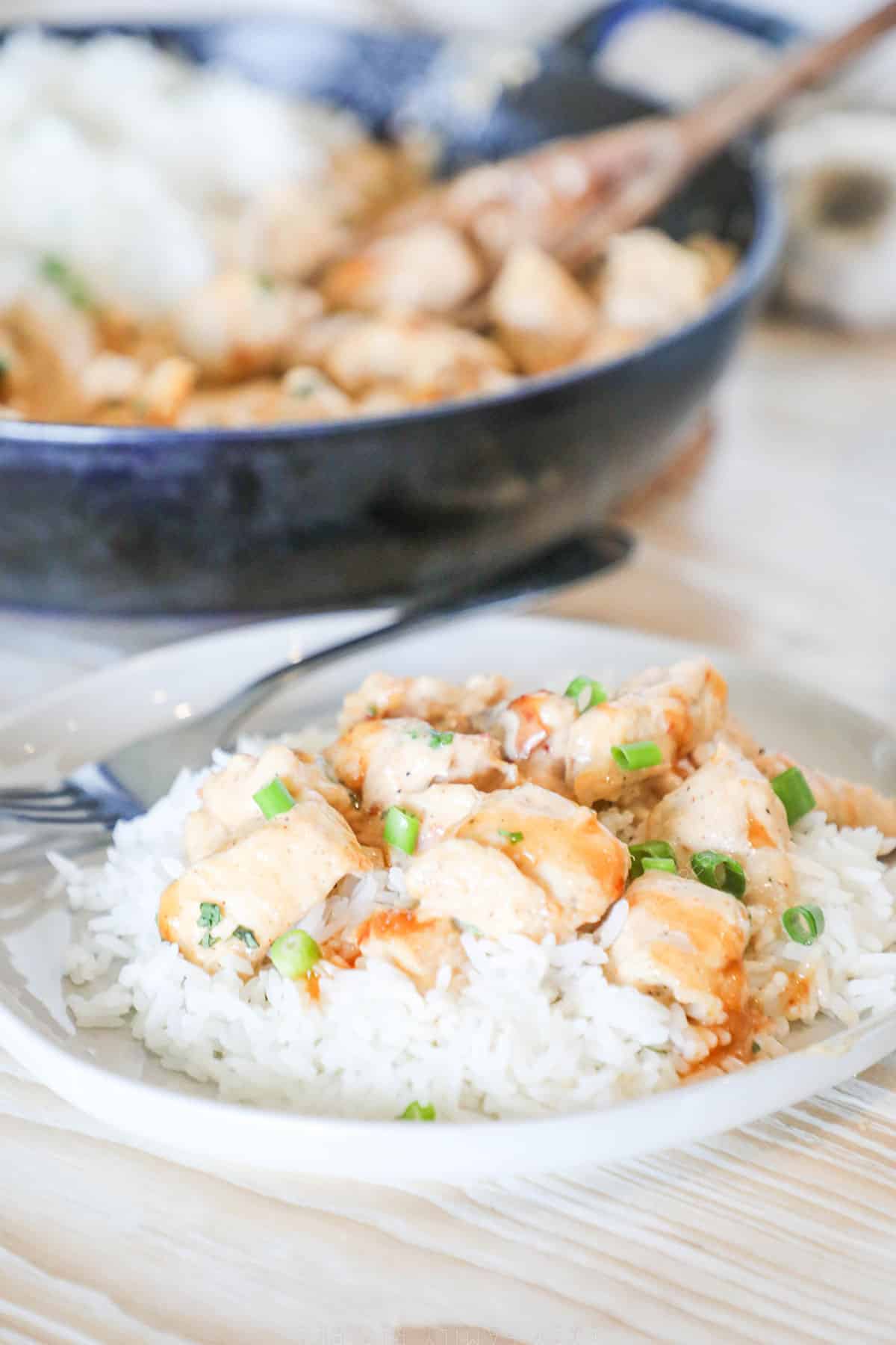 Bang bang chicken over rice on a white plate with a skillet full of chicken in the background.